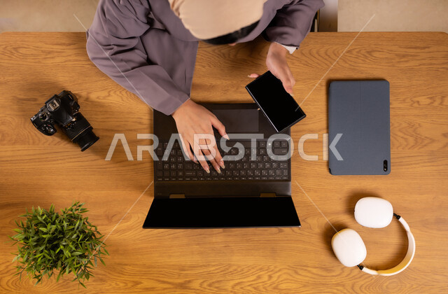 Using modern and advanced technologies, vertical overhead image of a Saudi Arabian Gulf woman holding a mobile phone in her hand using a laptop, integrating technology and technology into work, training via the Internet, editing shots and images via laptop applications, displaying a blank black screen
