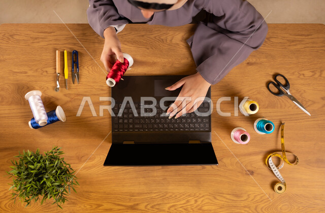 استخدام التكنولوجيا والتقنيات الحديثة، الإستفادة من المواقع الإلكترونية للتعلم والتدريب، الحصول على أدوات الخياطة أونلاين، صورة علوية عامودية لإمرأة عربية خليجية سعودية تستخدم الحاسوب المحمول تمسك بيدها الخيطان بجانبها مستلزمات الحياكة