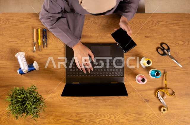 Using modern technology and techniques, benefiting from websites for learning and training, obtaining sewing tools online, vertical top view of a Saudi Arabian Gulf woman using a laptop holding a mobile phone next to her sewing supplies, displaying a blank black screen