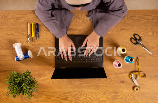 Using modern technology and techniques, taking advantage of websites for learning and training, obtaining sewing tools online, vertical top view of a Saudi Arabian Gulf woman using a laptop and next to her sewing supplies