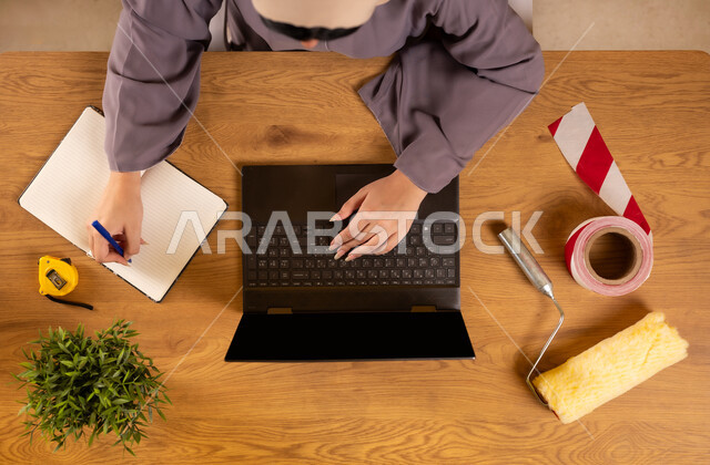 الاهتمام بالتجديد والتطور، الحصول على أدوات الدهان عبر الإنترنت، سهولة شراء مستلزمات المهن من خلال الاونلاين، استخدام التكنولوجيا والتقنيات الحديثة والمتطورة، صورة علوية عامودية لإمرأة عربية خليجية سعودية تستخدم الحاسوب وتقوم بتدوين الملاحظات على الدفتر