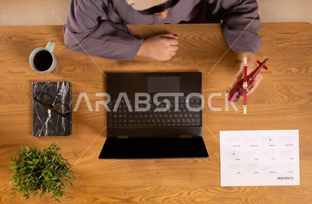 الاستعداد لقضاء رحلة صيفية ممتعة، مفهوم السياحة والقيام بالجولات الترفيهية، صورة علوية عامودية لإمرأة عربية خليجية سعودية تستخدم جهاز الحاسوب المحمول تحمل بيدها مجسم لطائرة، الحصول على المستلزمات اللازمة عن طريق الاونلاين، دمج التكنولوجيا في الحياة العملية