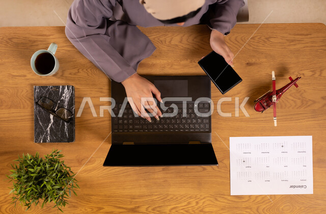 Concept of tourism and leisure tours, preparing for a fun summer trip, vertical overhead view of a Saudi Arabian Gulf woman using a phone and laptop, displaying a blank black screen, integrating technology into practical life