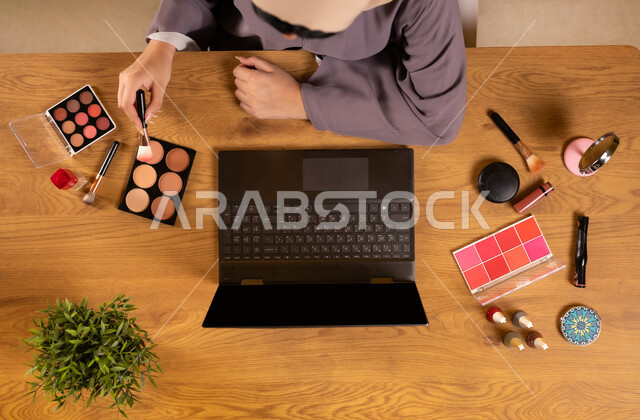 The concept of femininity and interest in beauty and appearance, modern women's cosmetics, marketing for makeup and makeup tutorials, integrating modern technology into daily life, vertical top view of a Saudi Arabian Gulf woman using cosmetic equipment next to her laptop