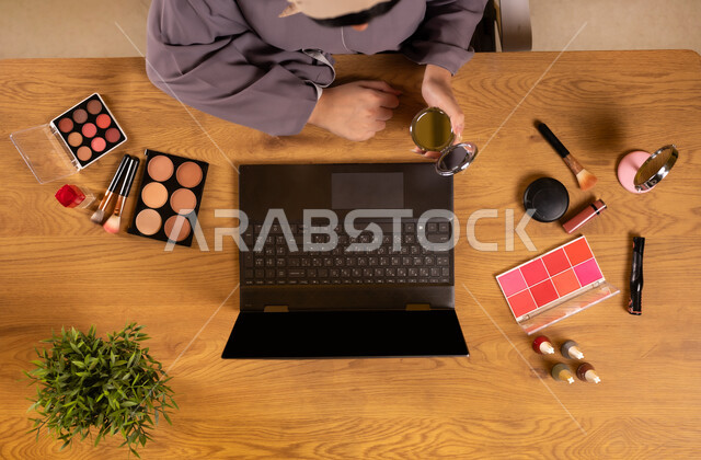 أدوات تجميل نسائية عصرية، مفهوم الأنوثة والاهتمام بالجمال والمظهر الخارجي، التسويق لمكياج وميكاب تتوريال، دمج التكنولوجيا الحديثة في الحياة اليومية، صورة علوية عامودية لإمرأة عربية خليجية سعودية تستخدم الحاسوب المحمول تحمل بيدها المرآة بجانبها معدات تجميلية