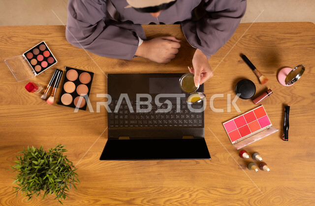 أدوات تجميل نسائية عصرية، مفهوم الأنوثة والاهتمام بالجمال والمظهر الخارجي، التسويق لمكياج وميكاب تتوريال، دمج التكنولوجيا الحديثة في الحياة اليومية، صورة علوية عامودية لإمرأة عربية خليجية سعودية تستخدم الحاسوب المحمول تحمل بيدها المرآة بجانبها معدات تجميلية
