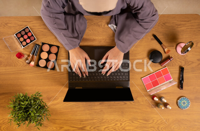 التسويق لمكياج وميكاب تتوريال، مفهوم الأنوثة والاهتمام بالجمال والمظهر الخارجي، أدوات تجميل نسائية عصرية، دمج التكنولوجيا الحديثة في الحياة اليومية، صورة علوية عامودية لإمرأة عربية خليجية سعودية تستخدم جهاز الحاسوب المحمول بجانبها المعدات التجميلية