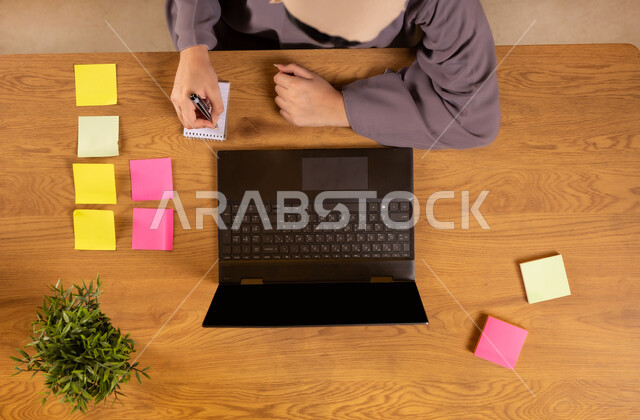 تدوين الأفكار وتسجيل المعلومات، تثبيت أوراق الملاحظات التذكارية الملونة، استخدام الأجهزة التقنية الحديثة في الأعمال المنزلية، انجاز مهام العمل عن بعد، صورة علوية عامودية لإمرأة عربية خليجية سعودية تستخدم جهاز الحاسوب المحمول تقوم بالكتابة على الدفتر