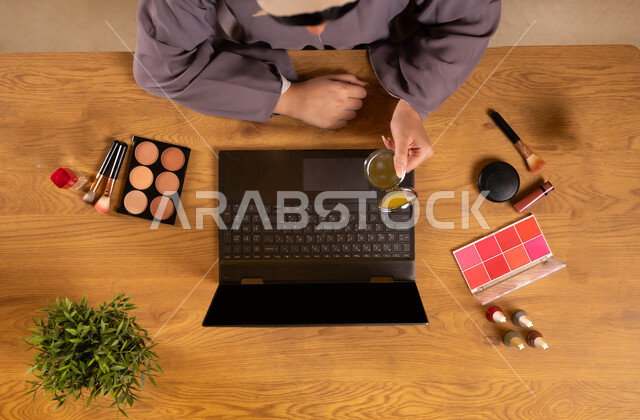 أدوات تجميل نسائية عصرية، مفهوم الأنوثة والاهتمام بالجمال والمظهر الخارجي، التسويق لمكياج وميكاب تتوريال، دمج التكنولوجيا الحديثة في الحياة اليومية، صورة علوية عامودية لإمرأة عربية خليجية سعودية تستخدم الحاسوب المحمول تحمل بيدها المرآة بجانبها معدات تجميلية