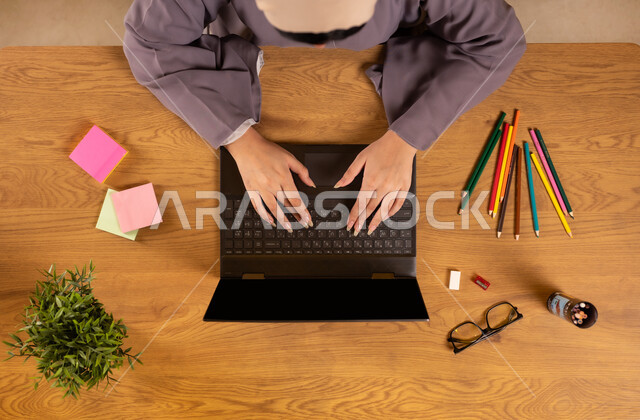 انجاز مهام العمل عن بعد، تدوين الأفكار وتسجيل المعلومات، تثبيت أوراق الملاحظات التذكارية الملونة، استخدام الأجهزة التقنية الحديثة في الأعمال المنزلية، صورة علوية عامودية لإمرأة عربية خليجية سعودية تستخدم الحاسوب المحمول وبجانبها الألوان والنظارات الطبية