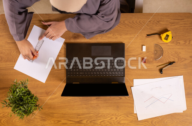 Integrating technology into scientific life, using modern and advanced technologies, learning arithmetic and mathematics through websites, vertical top view of a Saudi Arabian Gulf woman using a computer drawing graphs