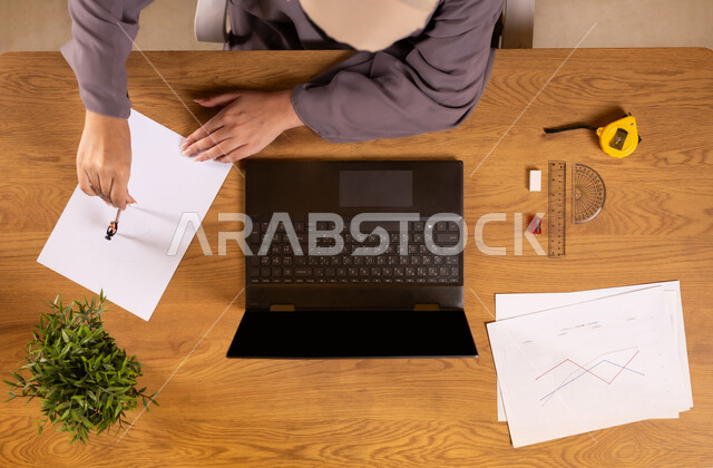 استخدام التقنيات الحديثة والمتطورة، تعلم الحساب والرياضيات من خلال المواقع الالكترونية، دمج التكنولوجيا في الحياة العلمية، صورة علوية عامودية لإمرأة عربية خليجية سعودية تمسك بيدها الهاتف المحمول تستخدم جهاز الحاسوب تقوم بتدوين الملاحظات، رسم المخططات البيانية