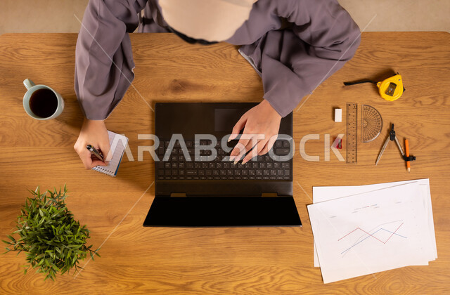 استخدام التقنيات الحديثة والمتطورة، تعلم الحساب والرياضيات من خلال المواقع الالكترونية، دمج التكنولوجيا في الحياة العلمية، صورة علوية عامودية لإمرأة عربية خليجية سعودية تمسك بيدها الهاتف المحمول تستخدم جهاز الحاسوب تقوم بتدوين الملاحظات، رسم المخططات البيانية