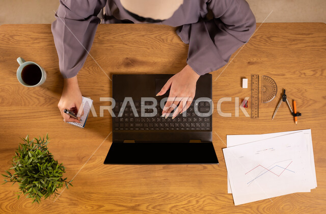 استخدام التقنيات الحديثة والمتطورة، تعلم الحساب والرياضيات من خلال المواقع الالكترونية، دمج التكنولوجيا في الحياة العلمية، صورة علوية عامودية لإمرأة عربية خليجية سعودية تمسك بيدها الهاتف المحمول تستخدم جهاز الحاسوب تقوم بتدوين الملاحظات، رسم المخططات البيانية