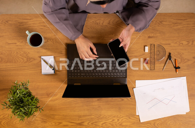 Learning arithmetic and mathematics through websites, integrating technology into scientific life, using modern and advanced technologies, vertical overhead image of a Saudi Arabian Gulf woman holding a mobile phone in her hand using a computer, drawing graphs, displaying a blank black screen