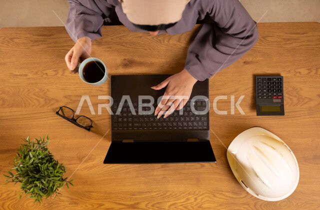 Integrating technology and modern devices into practical life, using calculator to perform calculations, supervising and auditing engineering projects and plans, vertical top view of a Saudi Arabian Gulf woman using a laptop, enjoying hot drinks