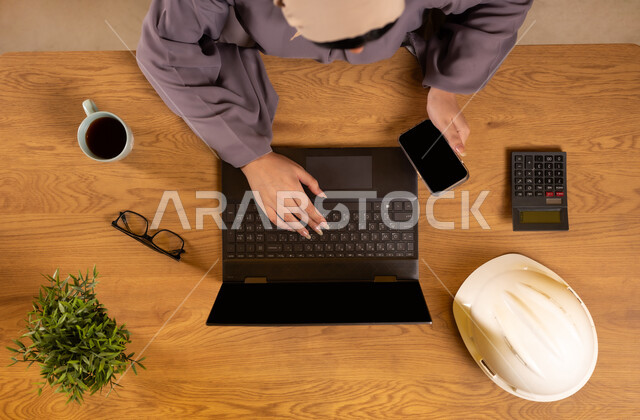 Integrating technology and modern devices into practical life, supervising and auditing engineering projects and plans, using calculator to perform calculations, vertical overhead image of a Saudi Arabian Gulf woman using a phone and laptop, displaying a blank black screen