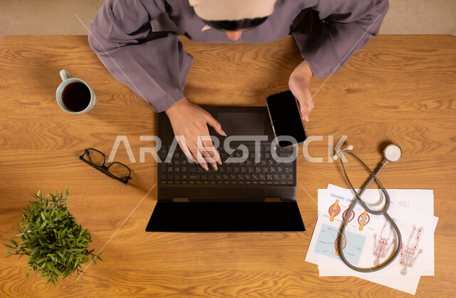 استخدام التقنيات الحديثة والمتطورة، مفهوم الطب والرعاية الصحية، دمج التكنولوجيا في الحياة العملية، صورة علوية عامودية لإمرأة عربية خليجية سعودية تمسك الهاتف بشاشة سوداء فارغة تعمل على جهاز الحاسوب المحمول