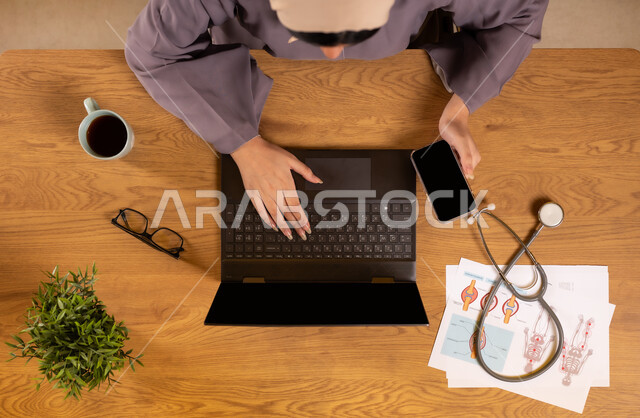 استخدام التقنيات الحديثة والمتطورة، مفهوم الطب والرعاية الصحية، دمج التكنولوجيا في الحياة العملية، صورة علوية عامودية لإمرأة عربية خليجية سعودية تمسك الهاتف بشاشة سوداء فارغة تعمل على جهاز الحاسوب المحمول