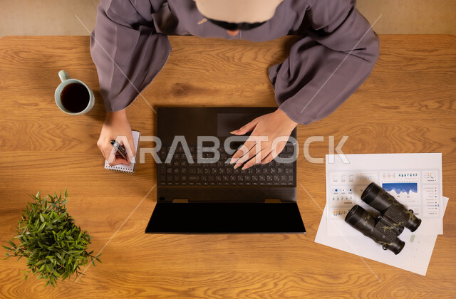 مفهوم البحث والاستكشاف، تدوين الملاحظات على الدفتر، مراقبة الإقتصاد والمعلومات المالية، دمج الأجهزة التقنية الحديثة بالحياة العملية، الإستثمار المالي الإلكتروني، صورة علوية عامودية لإمرأة عربية خليجية سعودية تقوم بدراسة وتحليل الإحصائيات عبر جهاز الحاسوب