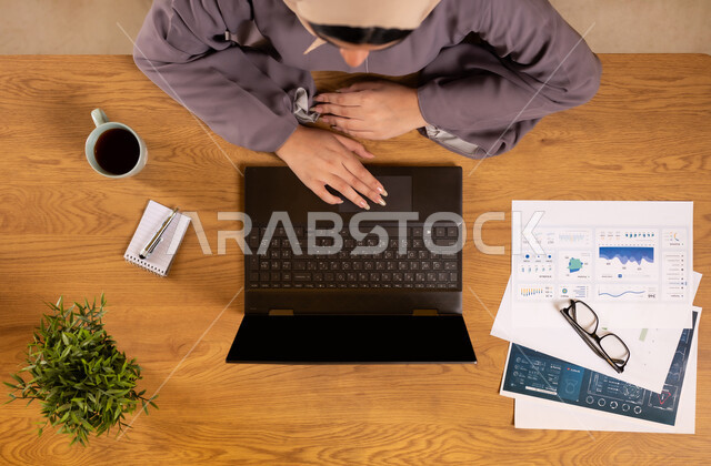 دمج الأجهزة التقنية الحديثة في الحياة العملية، متابعة مخطط الرسوم البيانية، الإستثمار المالي الإلكتروني، مراقبة الإقتصاد والمعلومات المالية، صورة علوية عامودية لإمرأة عربية خليجية سعودية تقوم بدراسة وتحليل الإحصائيات عبر جهاز الحاسوب المحمول، تناول المشروبات الساخنة