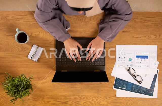 دمج الأجهزة التقنية الحديثة في الحياة العملية، متابعة مخطط الرسوم البيانية، الإستثمار المالي الإلكتروني، مراقبة الإقتصاد والمعلومات المالية، صورة علوية عامودية لإمرأة عربية خليجية سعودية تقوم بدراسة وتحليل الإحصائيات عبر جهاز الحاسوب المحمول، تناول المشروبات الساخنة