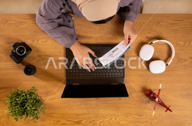 الاستعداد لقضاء رحلة صيفية ممتعة، مفهوم السياحة والقيام بالجولات الترفيهية، صورة علوية عامودية لإمرأة عربية خليجية سعودية تحمل بيدها تأشيرة السفر تقوم باستخدام جهاز الحاسوب المحمول، تثبيت الحجوزات اونلاين عبر تطبيقات وبرامج اللابتوب