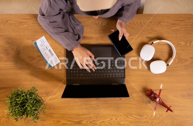 Integrating technology into practical life, preparing for a fun summer trip, the concept of tourism and leisure tours, a vertical aerial view of a Saudi Arabian Gulf woman using a laptop holding a phone with a blank black screen, making reservations online