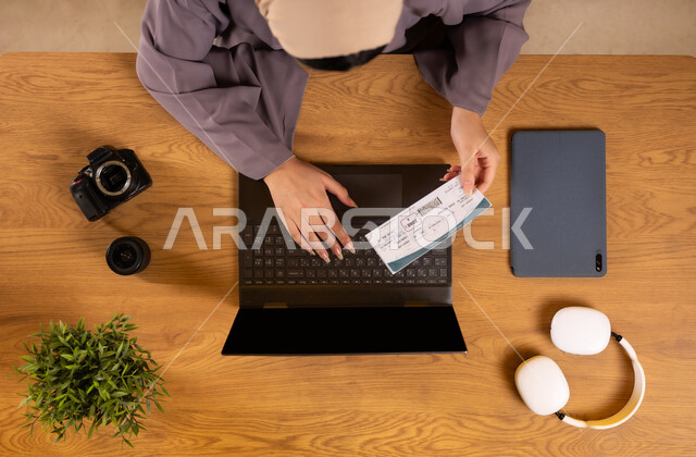 Preparing for a fun summer trip, the concept of tourism and recreational tours, vertical overhead view of a Saudi Arabian Gulf woman holding a travel visa using a laptop, making reservations online via laptop applications and programs