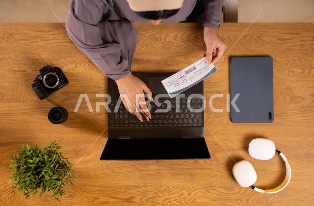 الاستعداد لقضاء رحلة صيفية ممتعة، مفهوم السياحة والقيام بالجولات الترفيهية، صورة علوية عامودية لإمرأة عربية خليجية سعودية تحمل بيدها تأشيرة السفر تقوم باستخدام جهاز الحاسوب المحمول، تثبيت الحجوزات اونلاين عبر تطبيقات وبرامج اللابتوب