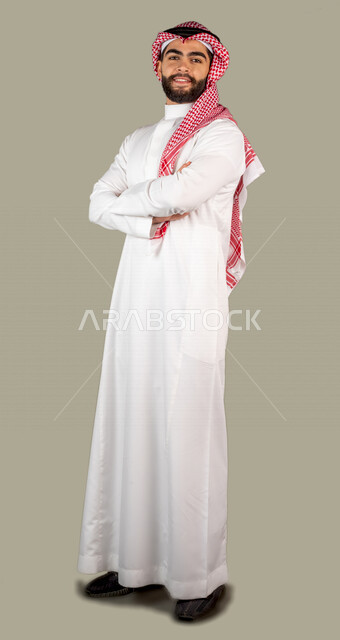 بورتريه لرجل عربي خليجي سعودي وسيم مكتف يديه يرتدي الزي السعودي التقليدي ، ايماءات و تعابير للوجه و اليدين ، خلفية رمادية ، كامل الجسم ، صورة بالطول الكامل
