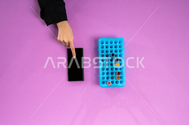 Blank black screen view, vertical overhead portrait of a Saudi Arabian Gulf woman holding a mobile phone and test tubes next to her, recording laboratory test results on the mobile phone, conducting scientific experiments and research via biotechnology, pink wrap