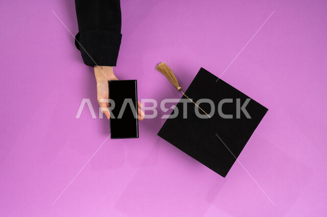 البيع والشراء عبر الانترنت، استخدام تطبيقات التسوق عن بعد، طلب مستلزمات التخرج أونلاين، عرض شاشة سوداء فارغة، بورتريه علوي عامودي ليد إمرأة عربية سعودية خليجية تعمل على جهاز الجوال وبجانبها قبعة تخرج، الاستفادة من التكنولوجيا الحديثة لمشروع التخرج، خلفية وردية
