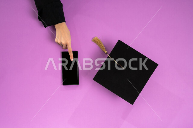 الاستفادة من التكنولوجيا الحديثة لمشروع التخرج، البيع والشراء عبر الانترنت، استخدام تطبيقات التسوق عن بعد، طلب مستلزمات التخرج أونلاين، عرض شاشة سوداء فارغة، بورتريه علوي عامودي ليد إمرأة عربية سعودية خليجية تعمل على جهاز الجوال وبجانبها قبعة تخرج، خلفية وردية