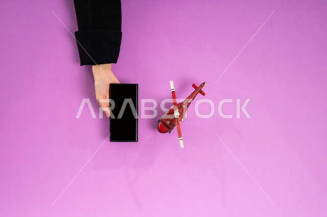 قطع تذاكر الطيران أونلاين، استخدام الانترنت للبحث عن رحلات جوية، مفهوم السياحة والقيام بالجولات الترفيهية، الاستعداد لقضاء رحلة صيفية ممتعة، عرض شاشة سوداء فارغة، بورتريه علوي عامودي ليد إمرأة عربية خليجية سعودية تستخدم الجوال وبجانبها مجسم طائرة، خلفية وردية