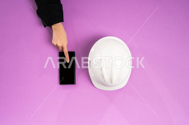 العمل في القطاع الهندسي، استخدام الأجهزة التقنية الحديثة بالأعمال الهندسية، عرض شاشة سوداء فارغة، بيع وشراء المستلزمات أونلاين، التدريب من خلال الانترنت، بورتريه علوي عامودي ليد إمرأة عربية سعودية خليجية تستخدم الجوال وبجانبها خوذة الحماية البيضاء، خلفية وردية