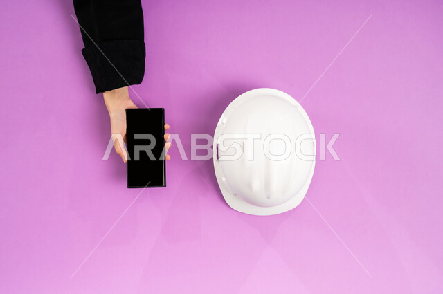 التدريب من خلال الانترنت، بورتريه علوي عامودي ليد إمرأة عربية سعودية خليجية تستخدم الجوال وبجانبها خوذة الحماية البيضاء، العمل في القطاع الهندسي، استخدام الأجهزة التقنية الحديثة بالأعمال الهندسية، عرض شاشة سوداء فارغة، بيع وشراء المستلزمات أونلاين، خلفية وردية