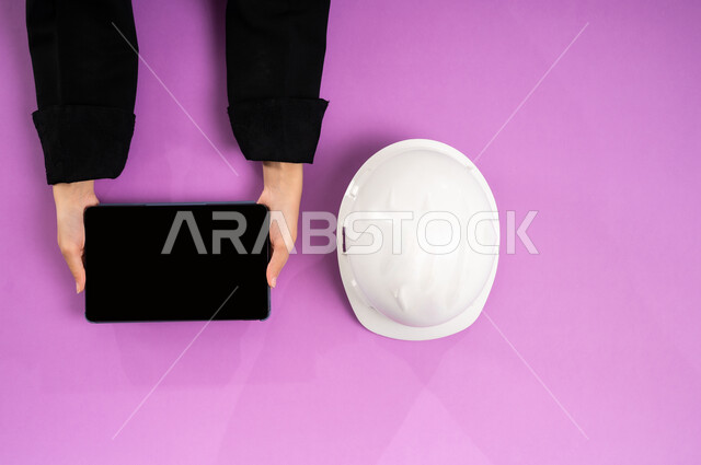 بيع وشراء المستلزمات الهندسية  أونلاين، التدريب من خلال الانترنت، بورتريه علوي عامودي ليد إمرأة عربية سعودية خليجية تستخدم التابليت وبجانبها خوذة الحماية البيضاء، العمل في القطاع الهندسي، استخدام الأجهزة التقنية الحديثة، عرض شاشة سوداء فارغة، خلفية وردية