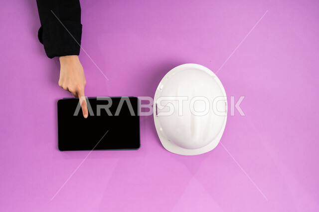 استخدام الأجهزة التقنية الحديثة، عرض شاشة سوداء فارغة، بيع وشراء المستلزمات الهندسية  أونلاين، التدريب من خلال الانترنت، بورتريه علوي عامودي ليد إمرأة عربية سعودية خليجية تستخدم التابليت وبجانبها خوذة الحماية البيضاء، العمل في القطاع الهندسي، خلفية وردية