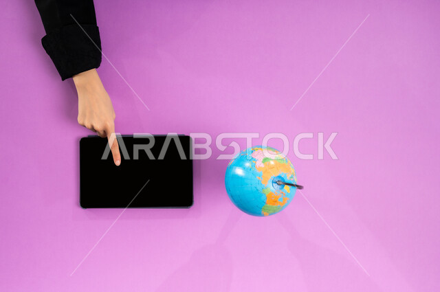 تحديد موقع الدول والبلدان العربية على خريطة العالم, البحث والاستكشاف حول الكون, مفهوم الرحلات والجولات السياحية, عرض شاشة سوداء فارغة، بورتريه علوي عامودي ليد إمرأة عربية سعودية خليجية تبحث عن مناطق للسفر عبر التابليت وبجانبها مجسم الكرة الأرضية، خلفية وردية