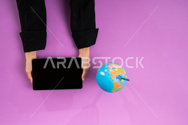 البحث والاستكشاف حول الكون, مفهوم الرحلات والجولات السياحية, عرض شاشة سوداء فارغة، بورتريه علوي عامودي ليد إمرأة عربية سعودية خليجية تبحث عن مناطق للسفر عبر التابليت وبجانبها مجسم الكرة الأرضية، تحديد موقع الدول والبلدان العربية على خريطة العالم, خلفية وردية
