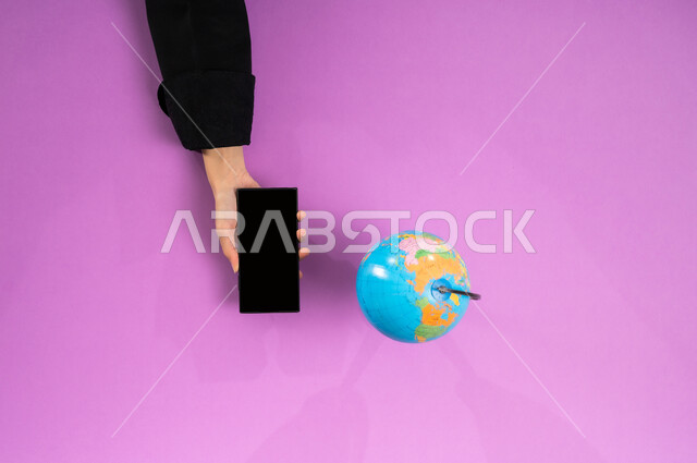 عرض شاشة سوداء فارغة، بورتريه علوي عامودي ليد إمرأة عربية سعودية خليجية تبحث عن مناطق للسفر عبر الجوال وبجانبها مجسم الكرة الأرضية، تحديد موقع الدول والبلدان العربية على خريطة العالم, البحث والاستكشاف حول الكون, مفهوم الرحلات والجولات السياحية, خلفية وردية