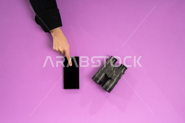 مفهوم النظرة المستقبلية، عرض شاشة سوداء فارغة، بورتريه علوي عامودي ليدين إمرأة عربية خليجية سعودية تستخدم الجوال وبجانبها المنظار ثنائي العينين، استعمال المنظار للكشف عن الاشياء البعيدة, حب الاستكشاف والمغامرة، التخطيط المستقبلي للأهداف والطموحات، خلفية وردية