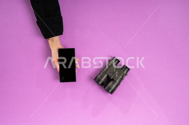 التخطيط المستقبلي للأهداف والطموحات، مفهوم النظرة المستقبلية، عرض شاشة سوداء فارغة، بورتريه علوي عامودي ليدين إمرأة عربية خليجية سعودية تستخدم الجوال وبجانبها المنظار ثنائي العينين، استعمال المنظار للكشف عن الاشياء البعيدة, حب الاستكشاف والمغامرة، خلفية وردية