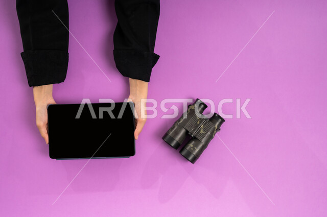 استعمال المنظار للكشف عن الاشياء البعيدة, حب الاستكشاف والمغامرة، التخطيط المستقبلي للأهداف والطموحات، مفهوم النظرة المستقبلية، عرض شاشة سوداء فارغة، بورتريه علوي عامودي ليدين إمرأة عربية خليجية سعودية تستخدم التابليت بجانبها المنظار ثنائي العينين، خلفية وردية