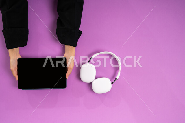 عرض شاشة سوداء فارغة، بورتريه علوي عامودي ليد إمرأة عربية خيجية سعودية تعمل على جهاز التابليت وبجانبها سماعة الرأس، الاستفادة من التكنولوجيا الحديثة، مفهوم الإصغاء والاستماع، استخدام تقنية الهيدفون، قضاء أوقات ممتعة بسماع الاغاني والمقاطع الصوتية، خلفية وردية