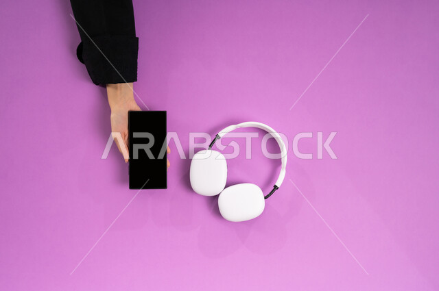 استخدام تقنية الهيدفون، قضاء أوقات ممتعة بسماع الاغاني والمقاطع الصوتية، عرض شاشة سوداء فارغة، بورتريه علوي عامودي ليد إمرأة عربية خيجية سعودية تعمل على الهاتف المحمول وبجانبها سماعة الرأس، الاستفادة من التكنولوجيا الحديثة، مفهوم الإصغاء والاستماع، خلفية وردية