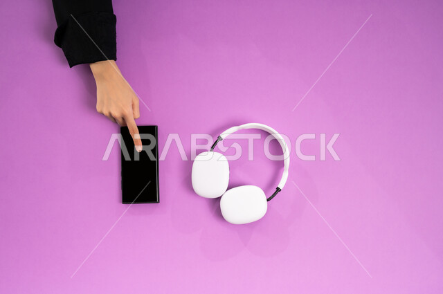 مفهوم الإصغاء والاستماع، إستخدام تقنية الهيدفون، قضاء أوقات ممتعة بسماع الاغاني والمقاطع الصوتية، عرض شاشة سوداء فارغة، بورتريه علوي عامودي ليد إمرأة عربية خيجية سعودية تعمل على الهاتف المحمول وبجانبها سماعة الرأس، الاستفادة من التكنولوجيا الحديثة، خلفية وردية