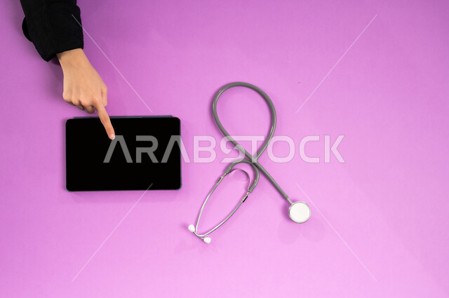 التسويق للأدوات الحديثة، بورتريه علوي عامودي ليدين إمرأة عربية سعودية خليجية تقوم بإجراء البحوث والدراسات عبر جهاز التابلت وبجانبها السماعة الطبية، عرض شاشة سوداء فارغة، طلب استشارة صحية أونلاين، مفهوم الطب والرعاية الصحية، الخدمات والأجهزة الطبية، خلفية وردية