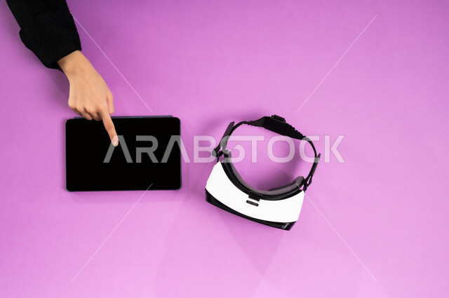 استخدام الأجهزة التقنية الحديثة, محاكاة العالم الافتراضي، التطور التكنولوجي في المملكة، الاستمتاع في العاب الفيديو، عرض شاشة سوداء فارغة، بورتريه علوي عامودي ليدين إمرأة عربية خليجية سعودية تعمل على التابليت وبجانبها نظارة الفي آر وقبضة التحكم، خلفية وردية